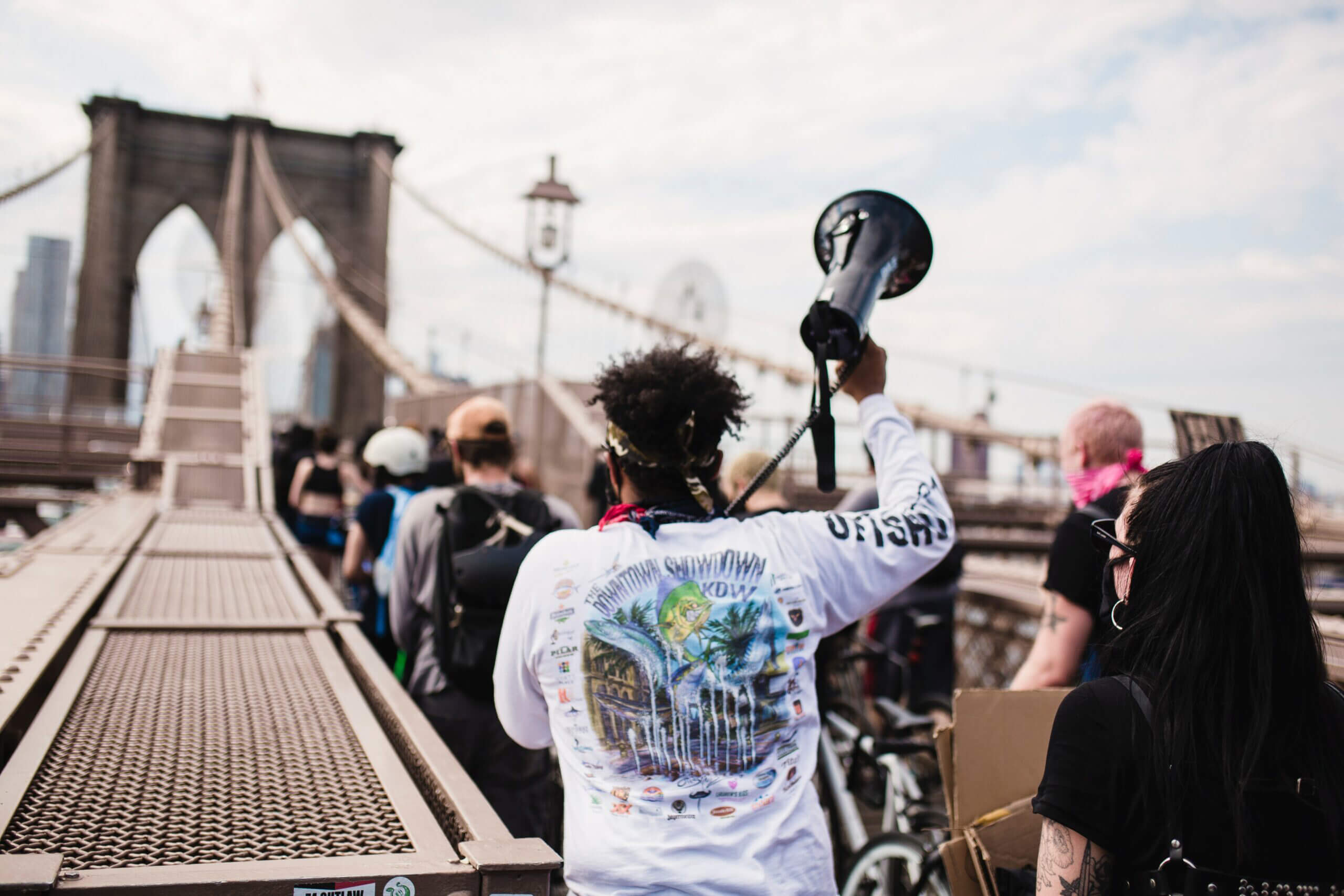 Black man holding a megaphone, at the centre of a protest, facing away from the camera, crossing the Brooklyn bridge