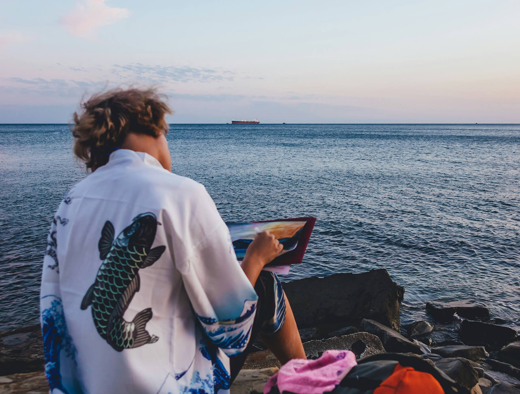 A woman sitting on a rock by the water, painting in a notebook