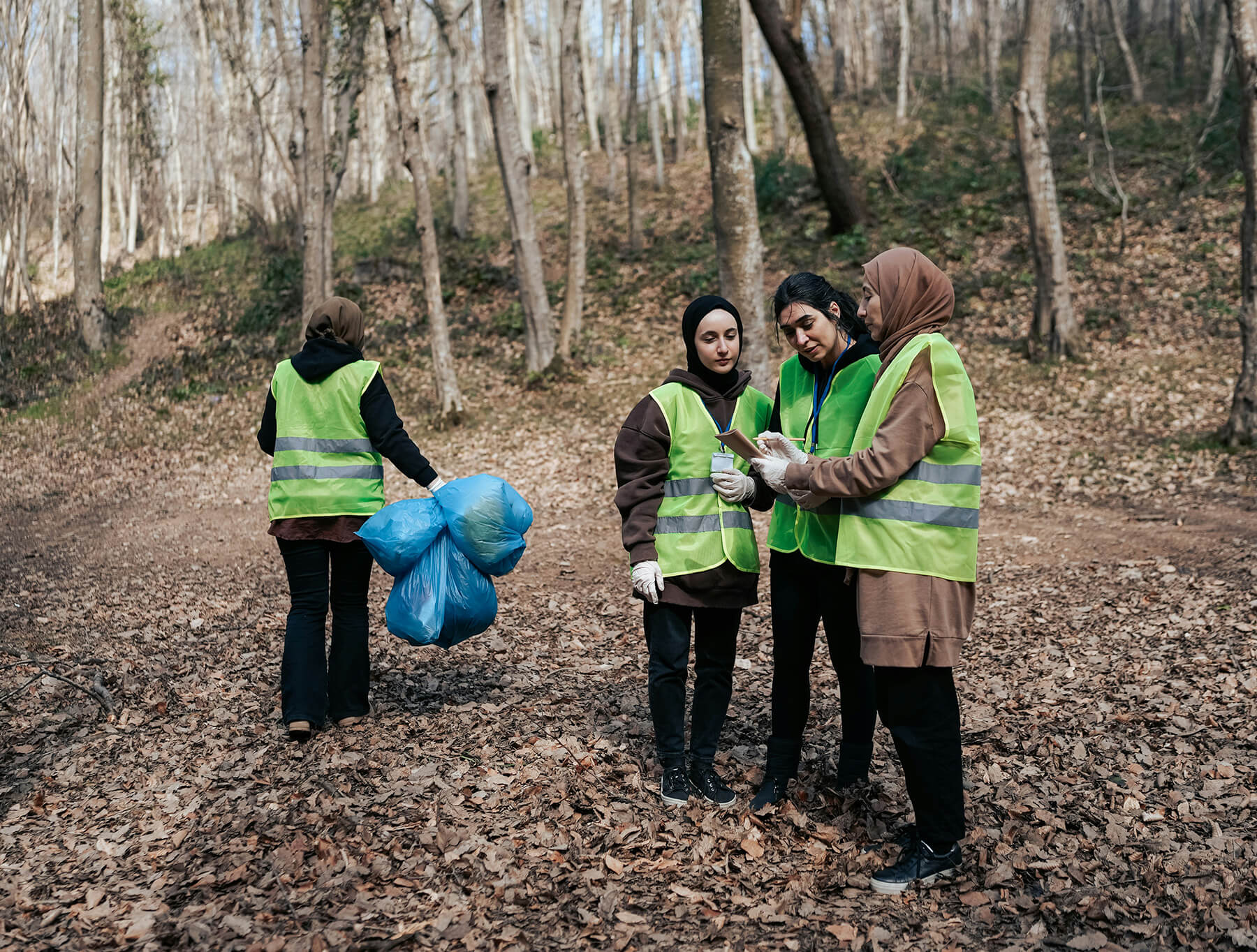 A group of women wearing hijabs and safety vests, picking up trash in the forest